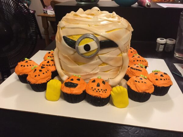 A cake decorated like a minion with pumpkins and cupcakes.