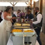 A bride and groom standing next to a buffet of food.