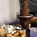 A chocolate fountain with fruit and snacks on a purple table.