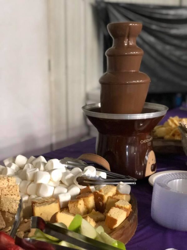 A chocolate fountain with fruit and snacks on a purple table.